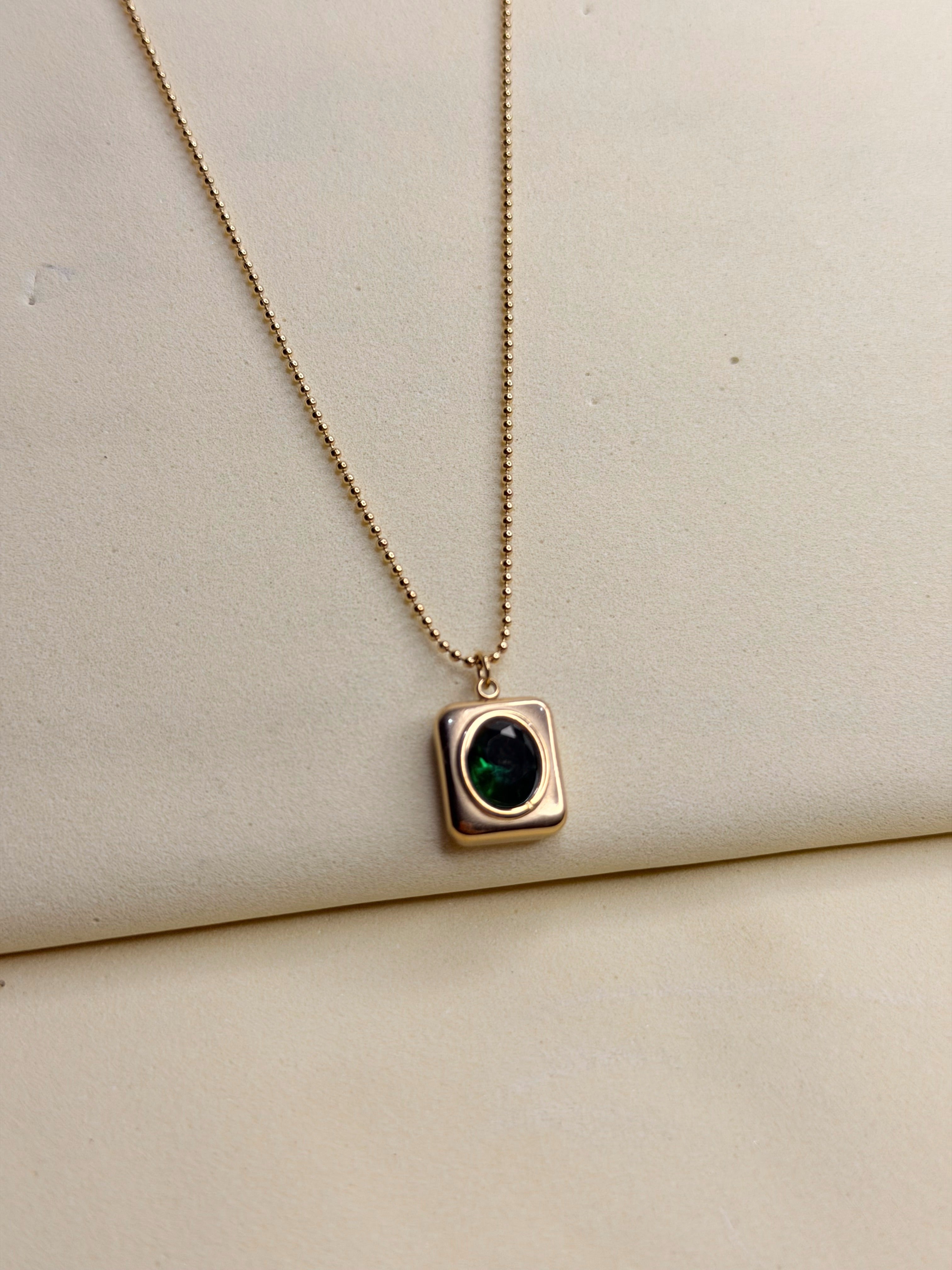 A gold-toned necklace with an emerald-green stone on a cream backdrop